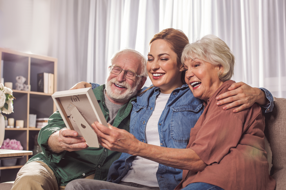 A woman sits on the couch between her elderly parents and they look at a photo frame together
