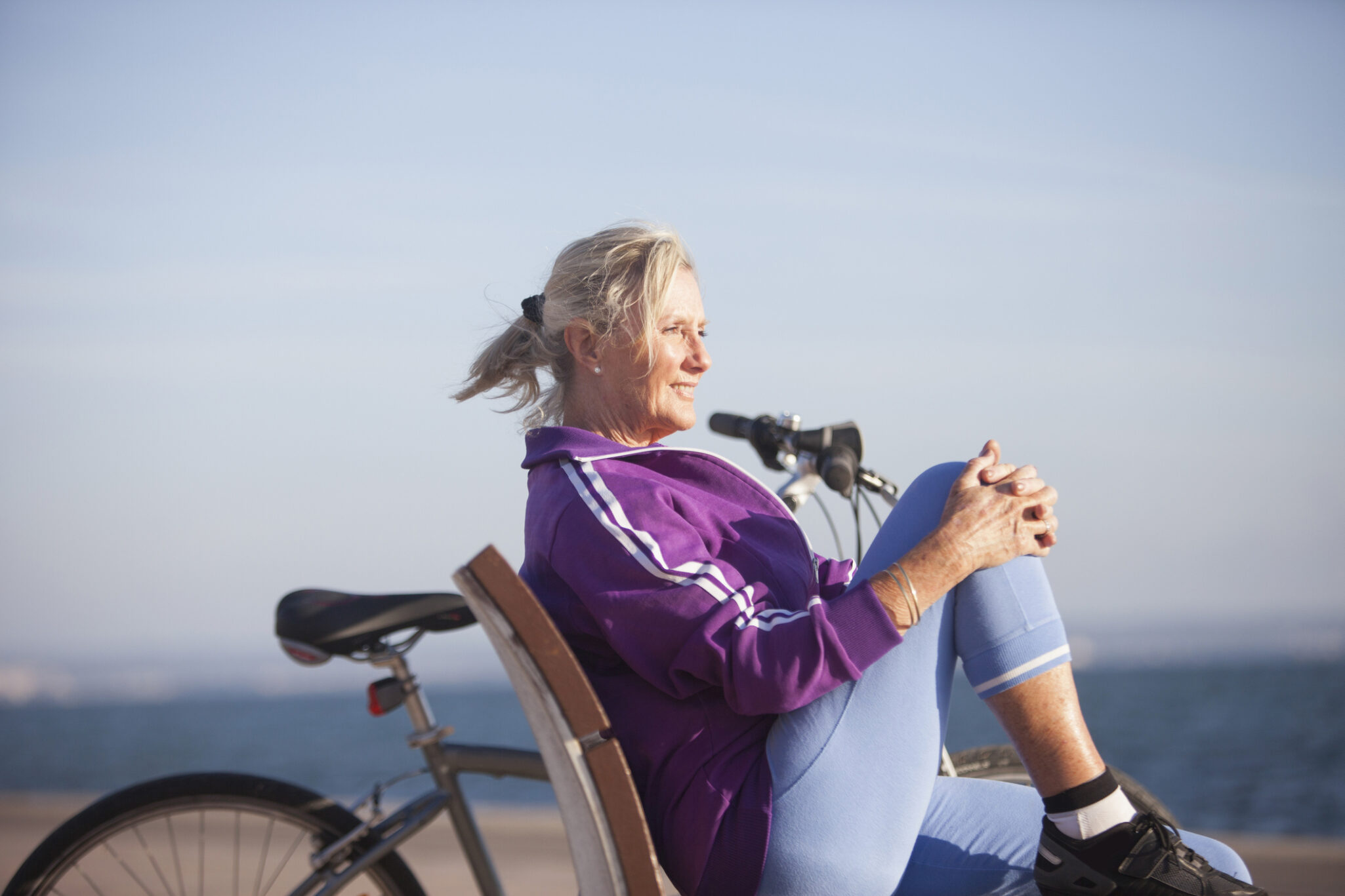 An older women takes a break from bike riding and sits on a beachside bench while holding one knee