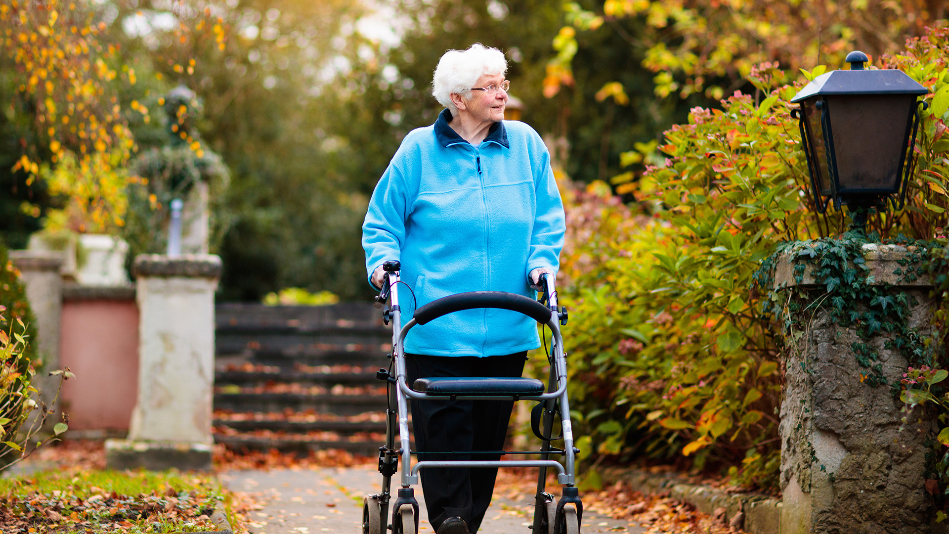 Happy senior handicapped lady with a walking disability enjoying a walk in an autumn park pushing her walker or wheel chair. Aid and support during retirement. Patient of nursing home or care center.